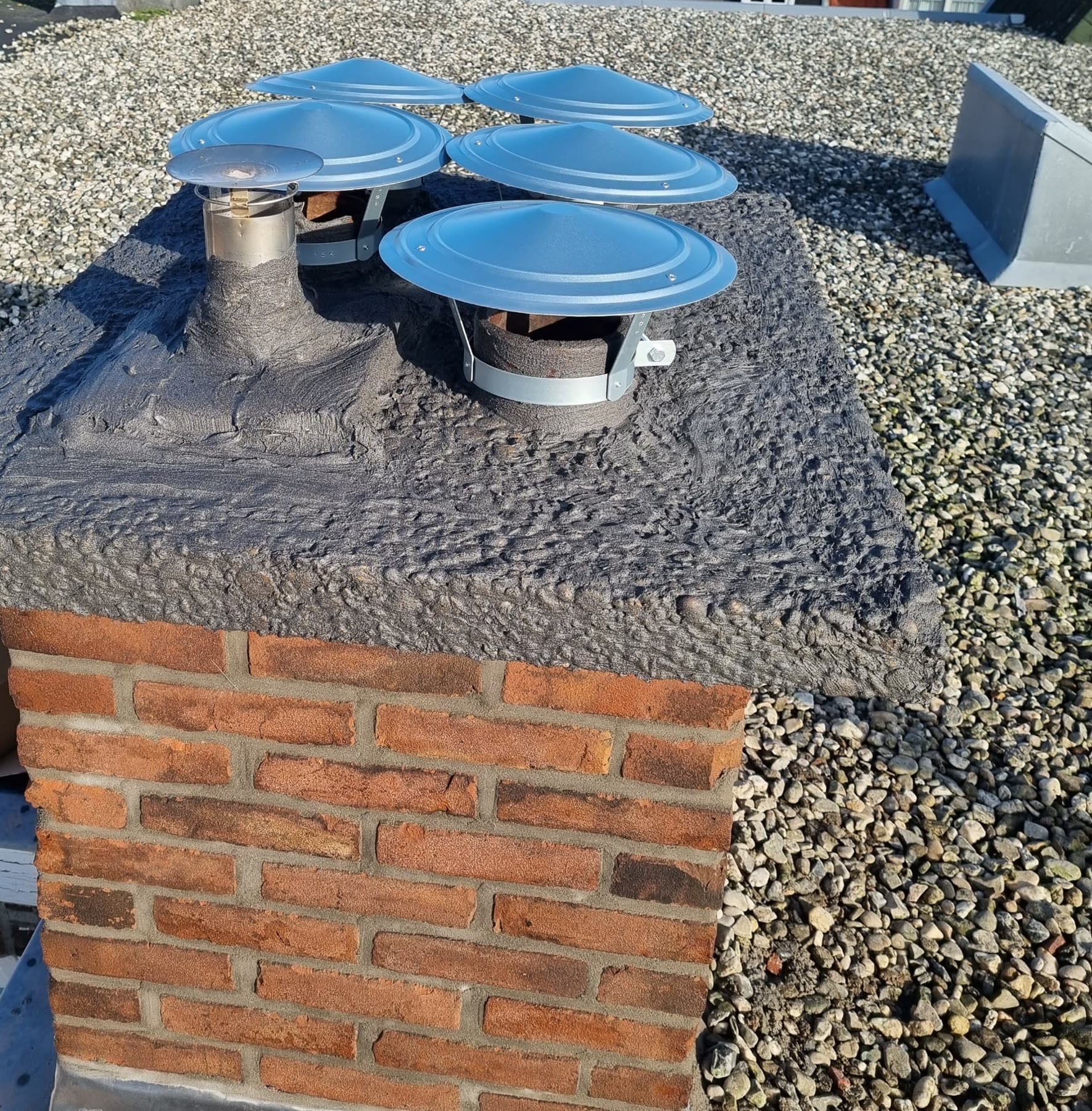 Een gemetselde schoorsteen op een grinddak in de zon: de afdekplaat is overgezet met een grijze, korrelige laag, met vijf nieuwe glimmende regenkappen en een rvs-rookkanaal erop; het metselwerk is felrood met lichte voegen
