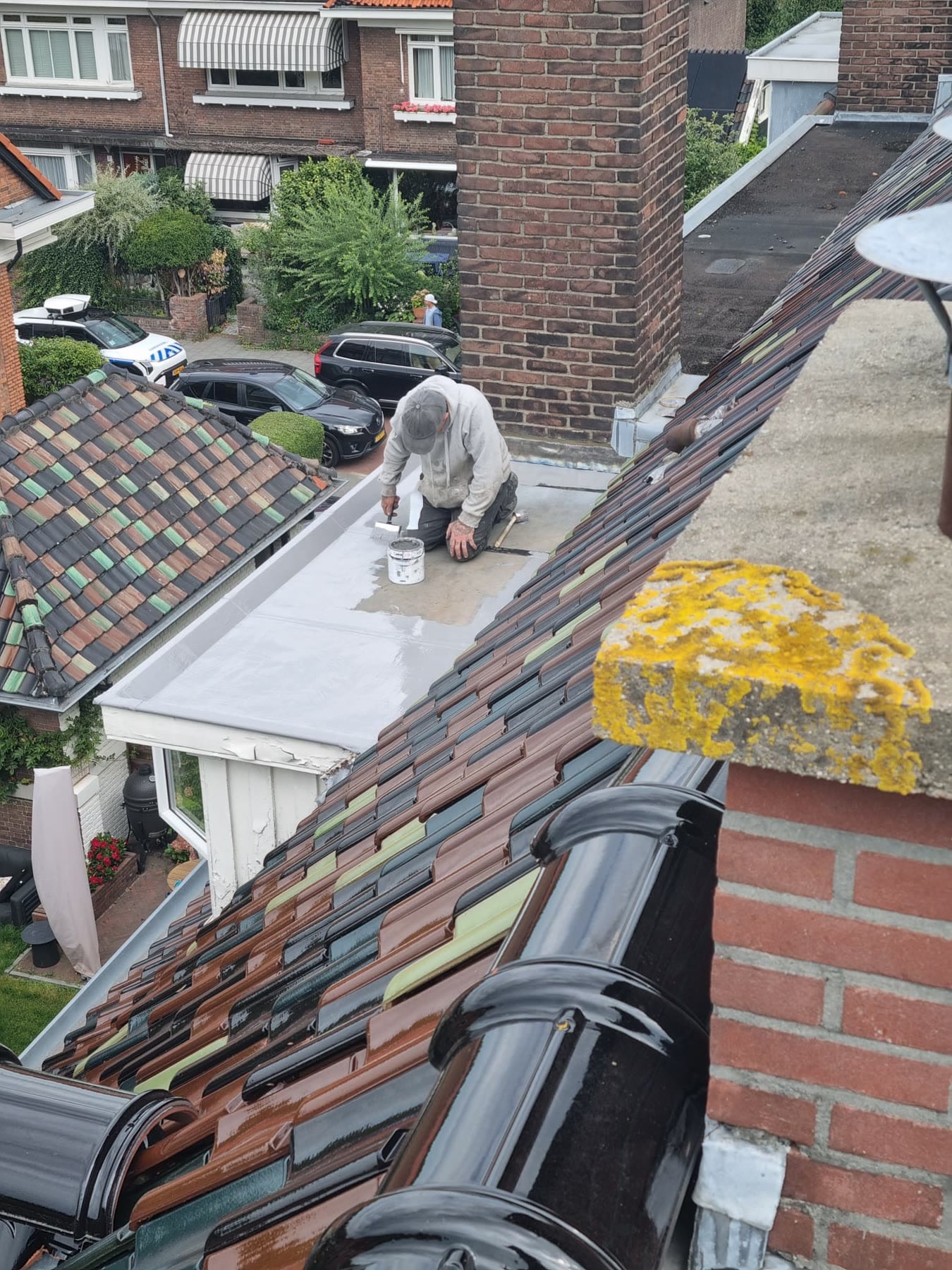 Een vakman knielt op het platte dak boven een witte erker en zet het met een kwast in met lichtgrijze coating uit een witte emmer; rechts vooraan een schoorsteenkap met gele korstmossen, beneden de straat met geparkeerde auto's